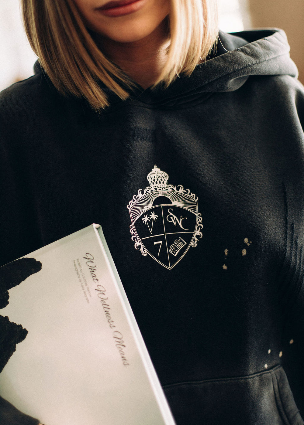 Model in the cream Safe Spaces Hoodie holding the What Wellness Means hardcover book from The Storyteller bundle